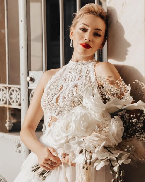 Bride in a Cycladic white boho wedding dress by Atelier Zolotas, featuring impressive crochet embroidery and holding a big white bouquet of roses